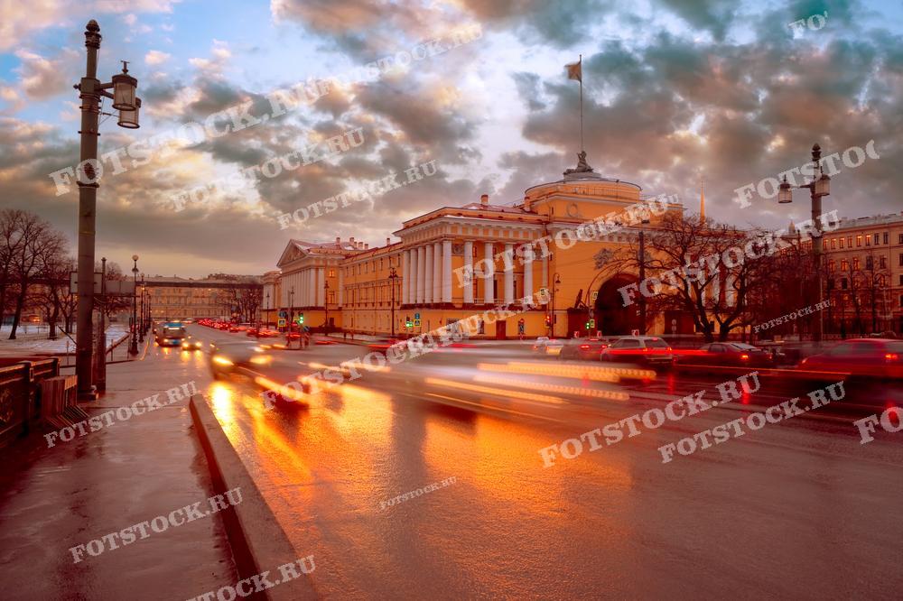 На фотографии изображено здание Адмиралтейства в Санкт-Петербурге на фоне закатного неба. Вечерние огни автомобилей создают красивые световые следы на улице. Закатные краски и облака придают картине особую атмосферу. Это место является одной из знаковых достопримечательностей города.